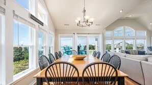A dining area with a long wooden table, black chairs, and a chandelier. Large windows offer views of the outdoors. - Destination-OBX Soundfront, 8 bdrm, 10 bth, elavator, pool, hot tub, game room (Nags Head)