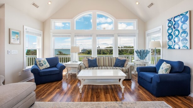 A spacious living room with a high ceiling, large windows, and a view of the outdoors. - Destination-OBX Soundfront, 8 bdrm, 10 bth, elavator, pool, hot tub, game room (Nags Head)