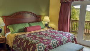 A bedroom with a large bed, green and red pillows, a patterned bedspread, a bedside lamp, a nightstand, and a view of a balcony with a wooden railing.
