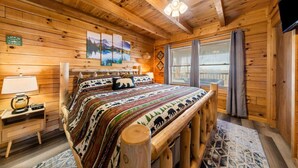 A wooden cabin bedroom with a bed, a nightstand, a lamp, a window with curtains, and a wall with a picture. - NEW! Updated Family Log Cabin+Hot Tub+Pool Table! (Sevierville)