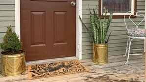 Una puerta principal con puerta marrón y ventana con el rótulo 'SWEET'. Hay dos macetas y una silla blanca en el porche.