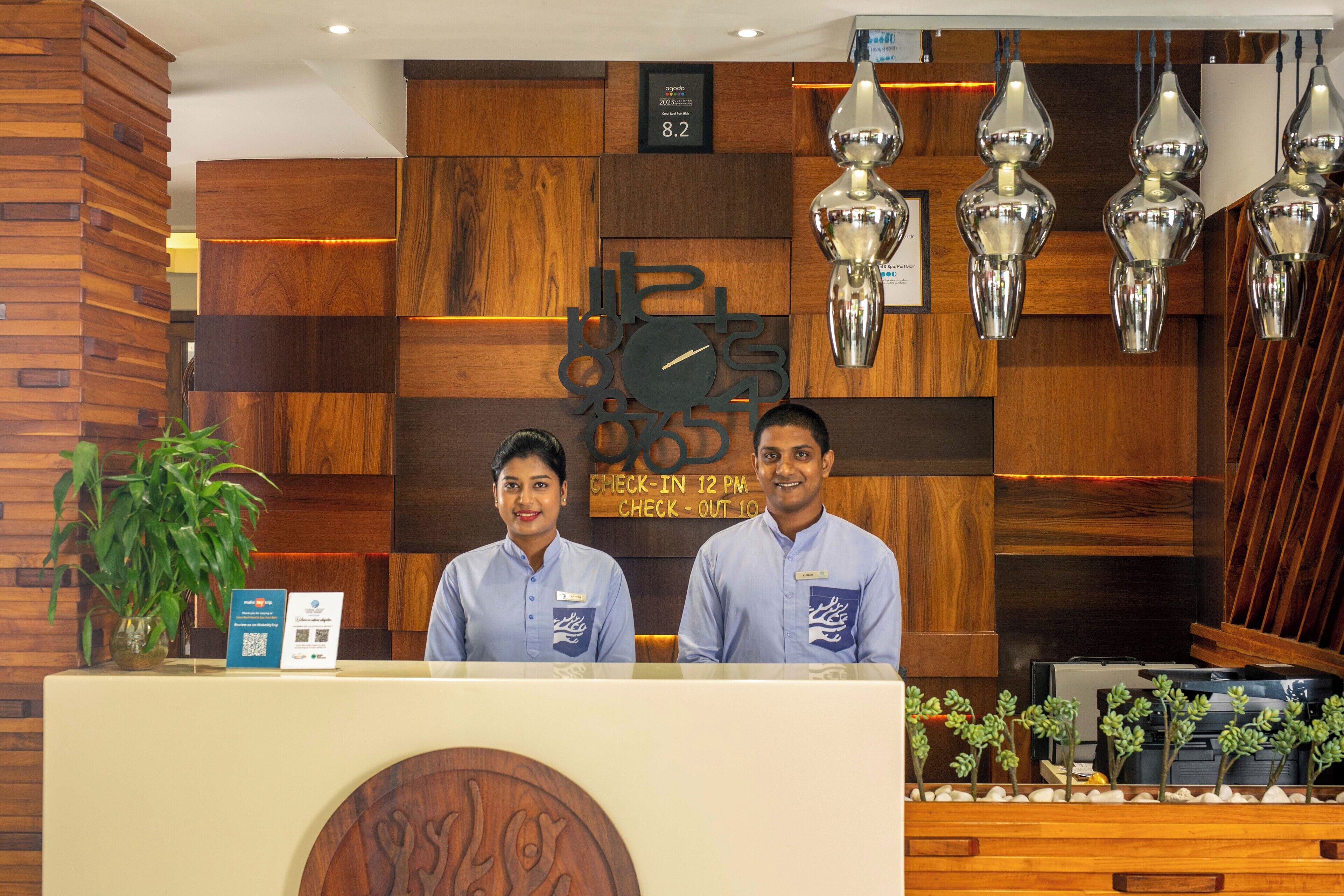 A reception desk with wooden paneling and a clock. There are two staff members behind the desk, and a plant to the left.