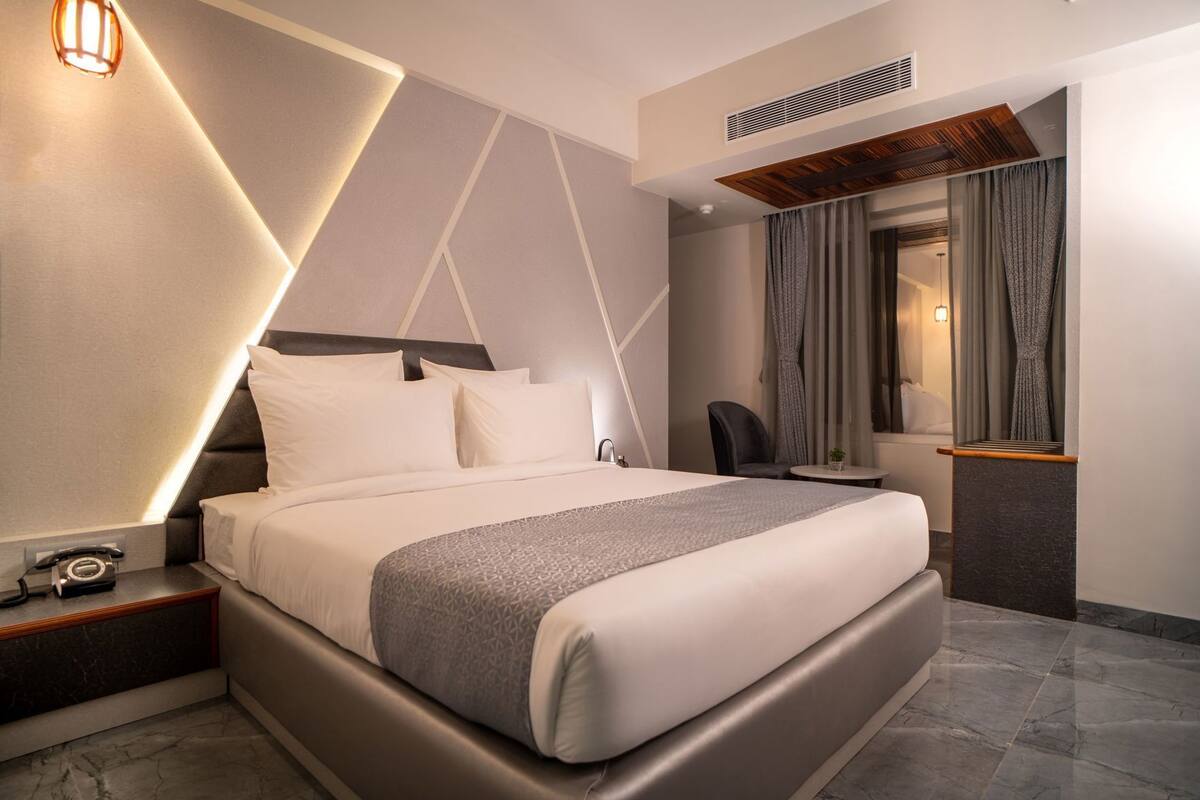 A modern hotel room with a large bed, a bedside table with a telephone, a small table with a plant, and a window with curtains.