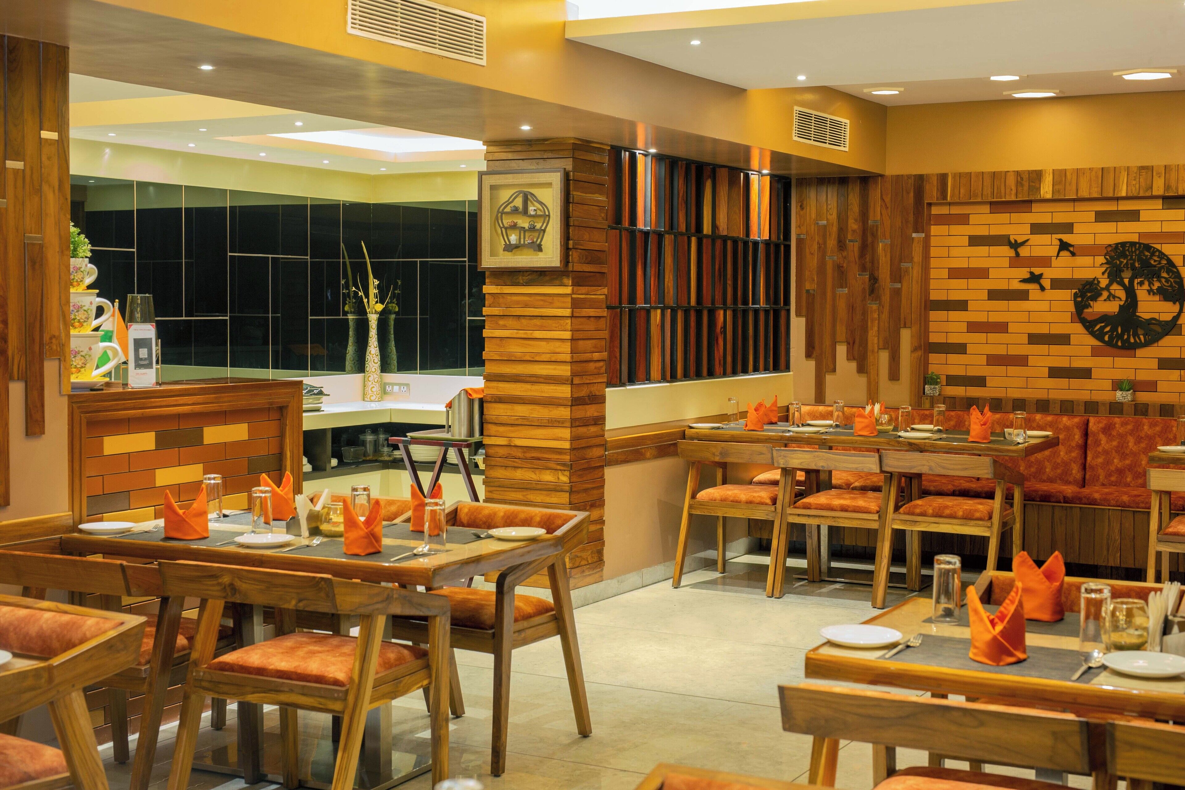A restaurant with wooden furniture and orange cushions. Tables are set with plates, glasses, and napkins. The walls have a wooden panel design and a mural of a tree.