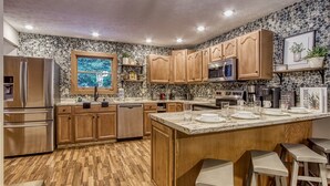 A modern kitchen with wooden cabinets, a large island with stools, a stone backsplash, and a window with a view of trees.