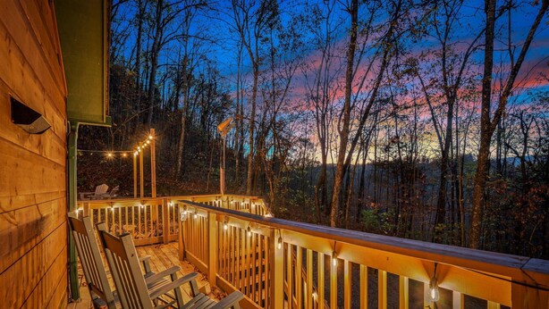 A wooden deck with string lights, wooden chairs, and a view of a forested area at dusk.