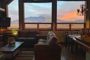 A cozy living room with a large window offering a mountain view. There is a sofa, a television, and a dining area with a table and chairs.