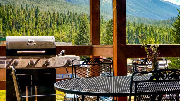A wooden deck with a grill, outdoor seating, and a view of a forested mountain range. - Luxury Yellowstone Chalet - Mountain Views with Sauna! (Cooke City-Silver Gate)