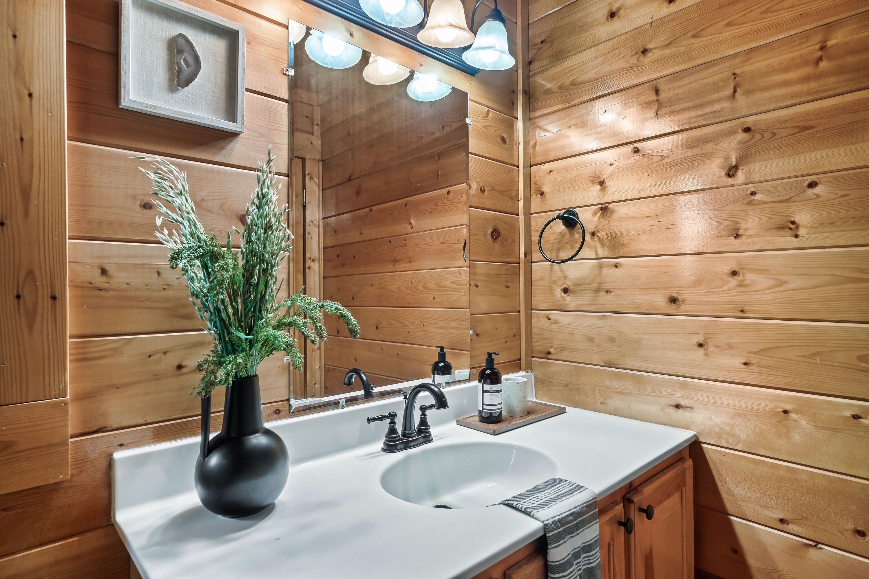 A wooden cabin-style bathroom with a white sink, black faucet, and a mirror above it. There is a black vase with green plants on the sink counter, and a wooden shelf with a white cup and a black bottle.
