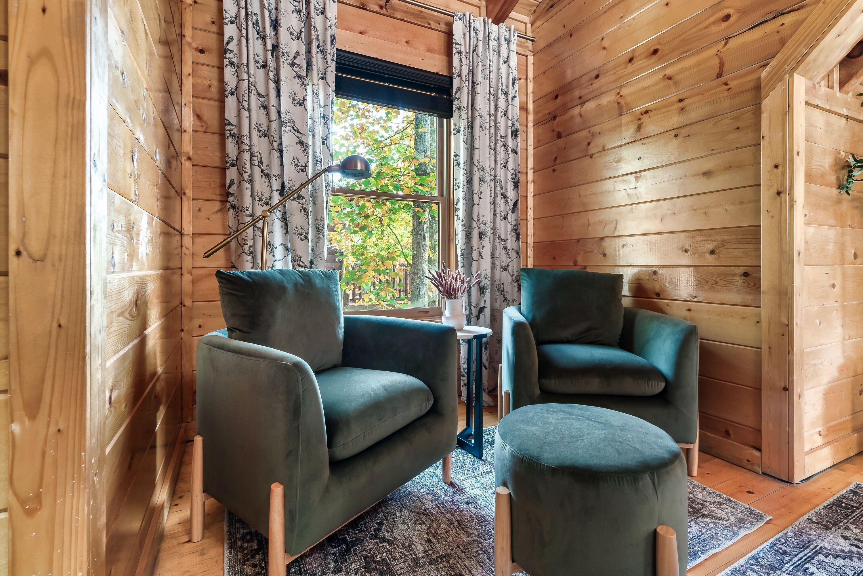 A cozy room with wooden walls, two green armchairs, a small table with a lamp, and a window with curtains.