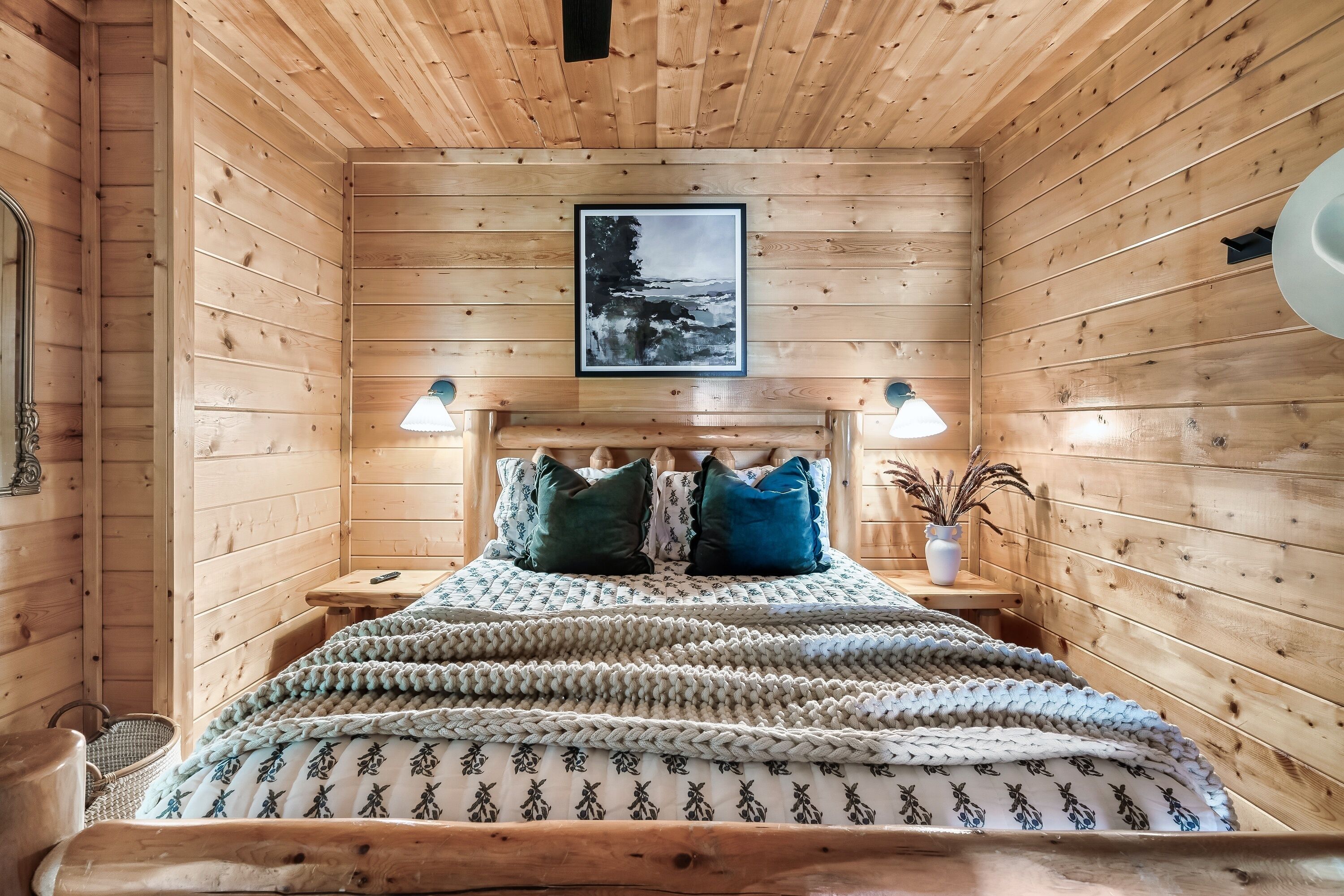 A cozy wooden cabin room with a bed, two bedside lamps, a framed picture on the wall, and a small table with a vase.