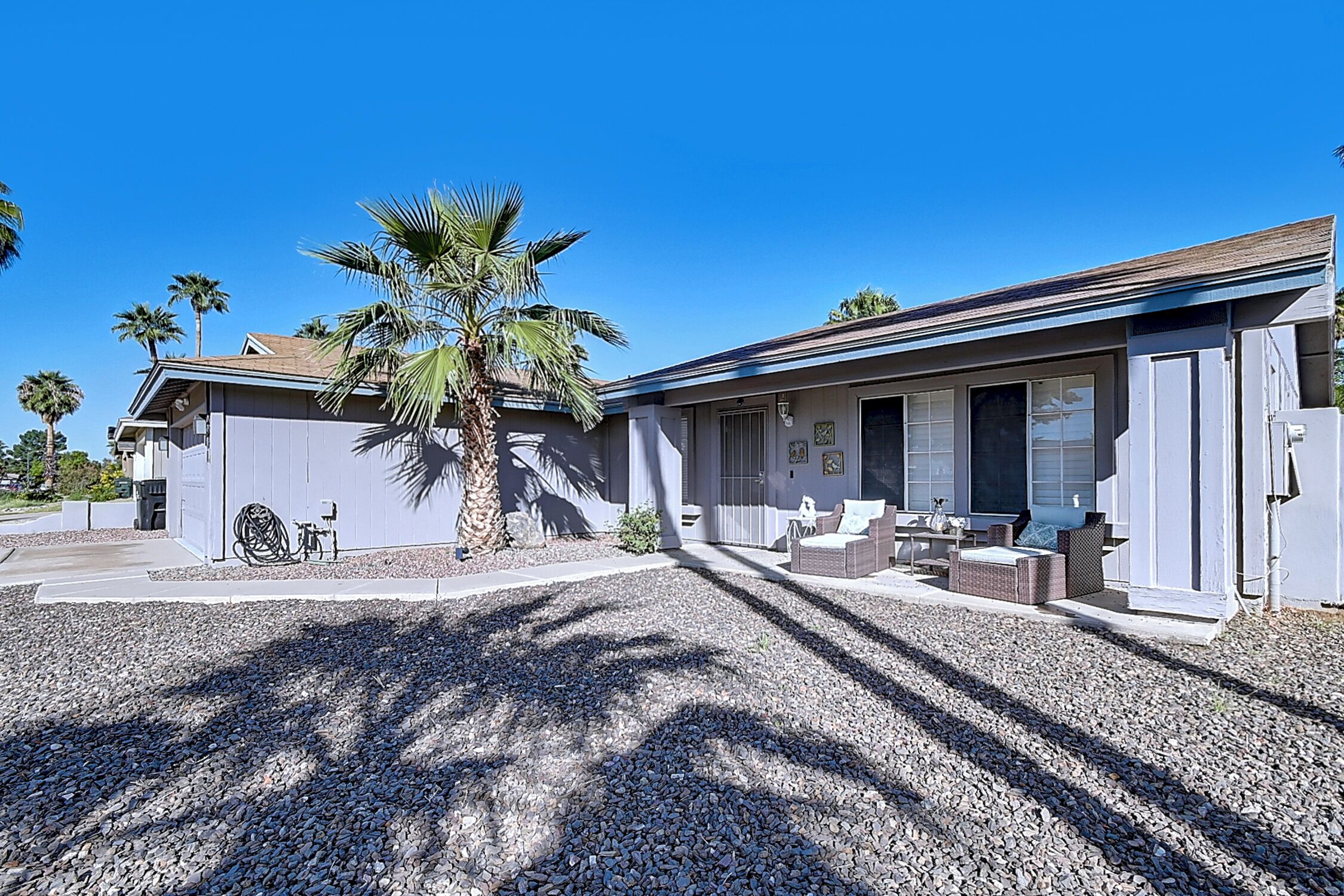 A two-story house with a covered patio, a palm tree, and a gravel driveway.
