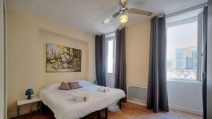 Une chambre à coucher comprenant un lit, une table de chevet, une lampe, un tableau, une fenêtre avec des rideaux et un ventilateur de plafond.
