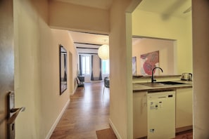 A hallway with wooden flooring, a dining area with chairs, a kitchen with a dishwasher, and a large window with curtains.