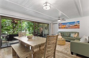A spacious living area with a wooden dining table, a green sofa, and a large window offering a view of lush greenery.