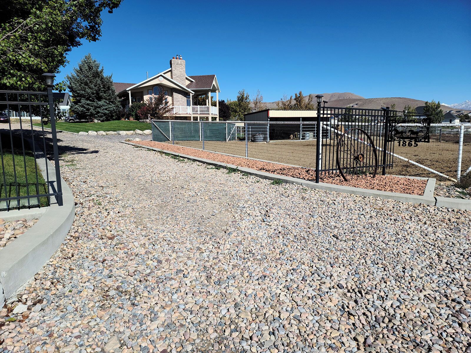 A gravel driveway leading to a house with a chimney, a fenced area, and a mountain in the background.