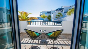 A balcony with two chairs and a small table, overlooking a white building with blue accents and palm trees.