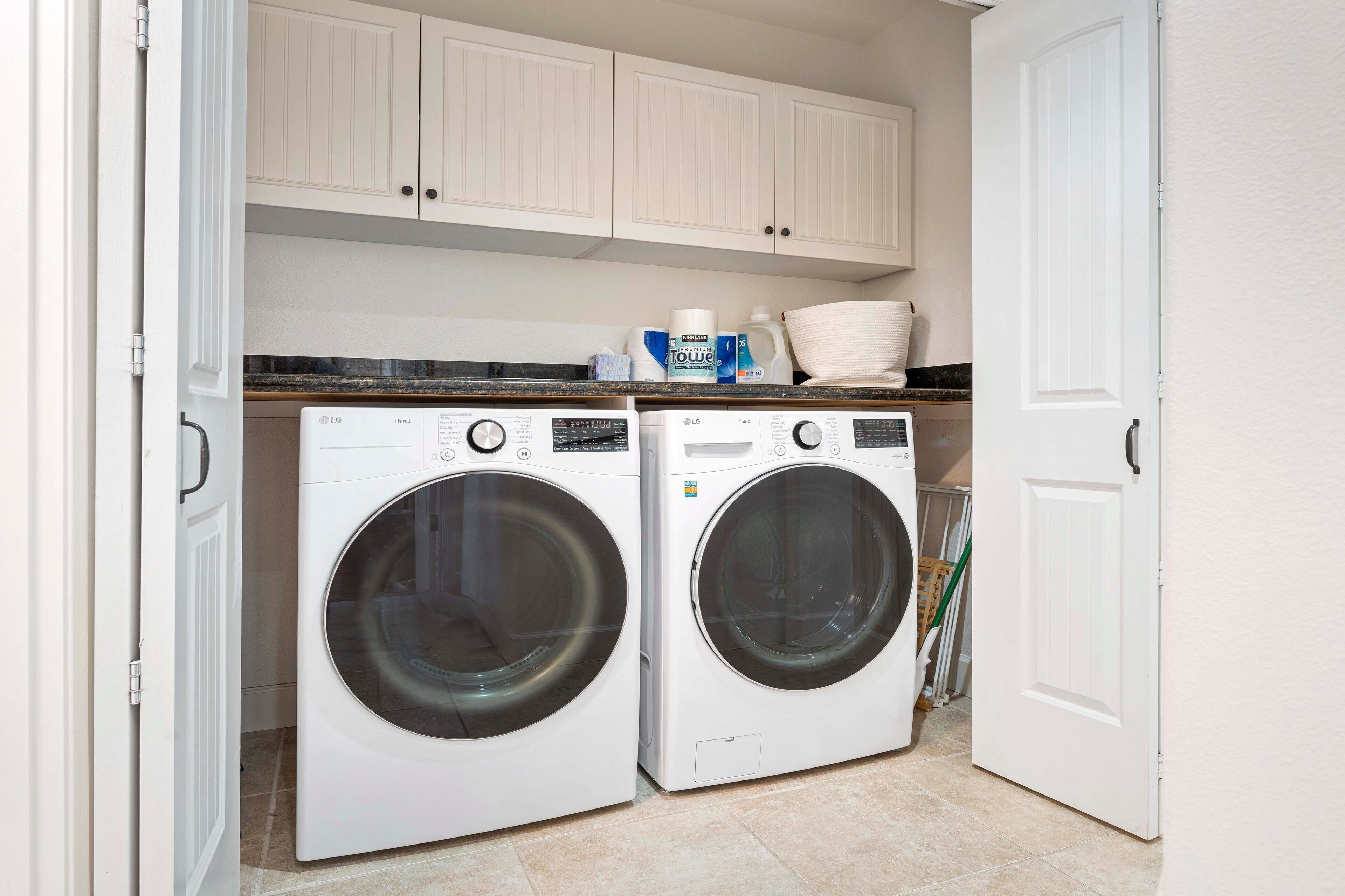 A laundry room with two white front-loading washing machines, a countertop with cleaning supplies, and white cabinets above.