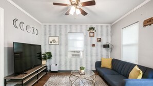 A living room with a TV on a wooden stand, a glass coffee table, a blue sofa with yellow cushions, a ceiling fan, and a wall with a moon phase decoration.