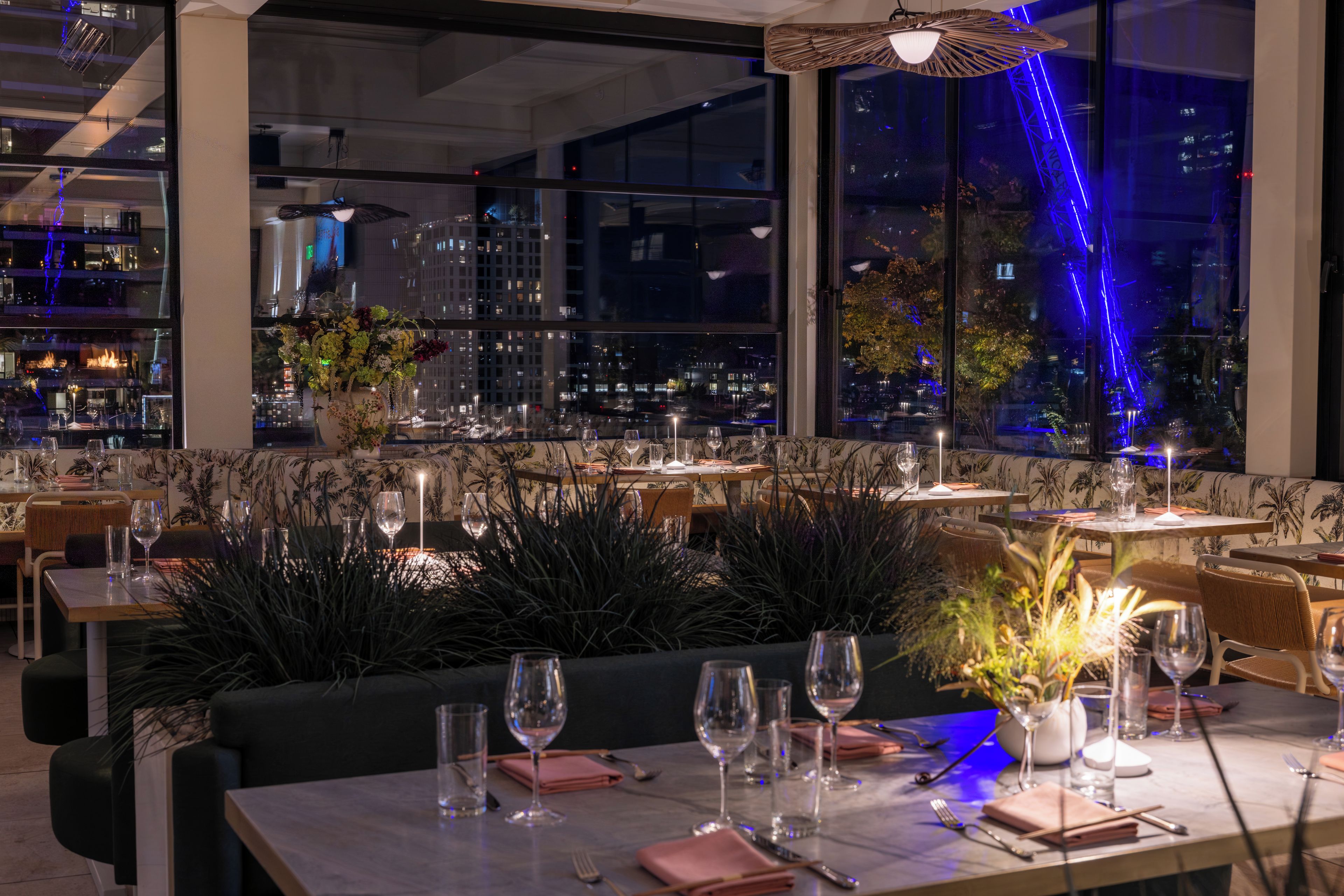Ein Restaurant mit gedeckten Tischen, großen Fenstern mit Blick auf die Stadt und Zimmerpflanzen.