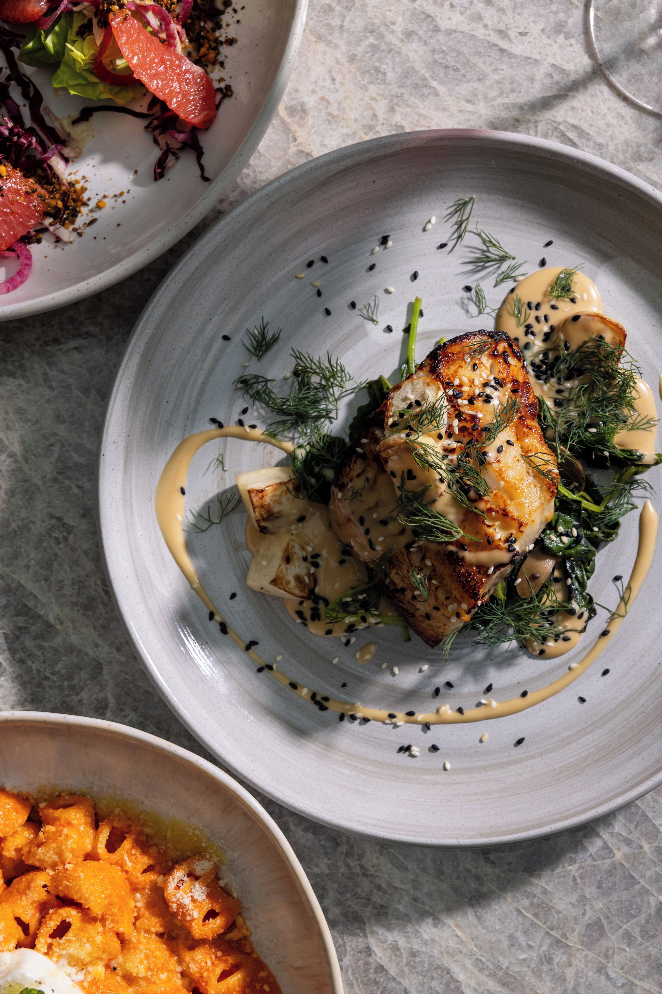 Un plato con un trozo de pescado a la parrilla, adornado con eneldo y semillas de sésamo, acompañado de una salsa cremosa y una guarnición de verduras.