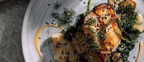 Un plato con un trozo de pescado a la parrilla, adornado con eneldo y semillas de sésamo, acompañado de una salsa cremosa y una guarnición de verduras.