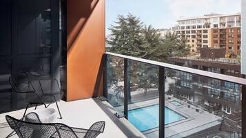 A balcony with two black wire mesh chairs, a glass railing, and a view of a swimming pool and buildings.