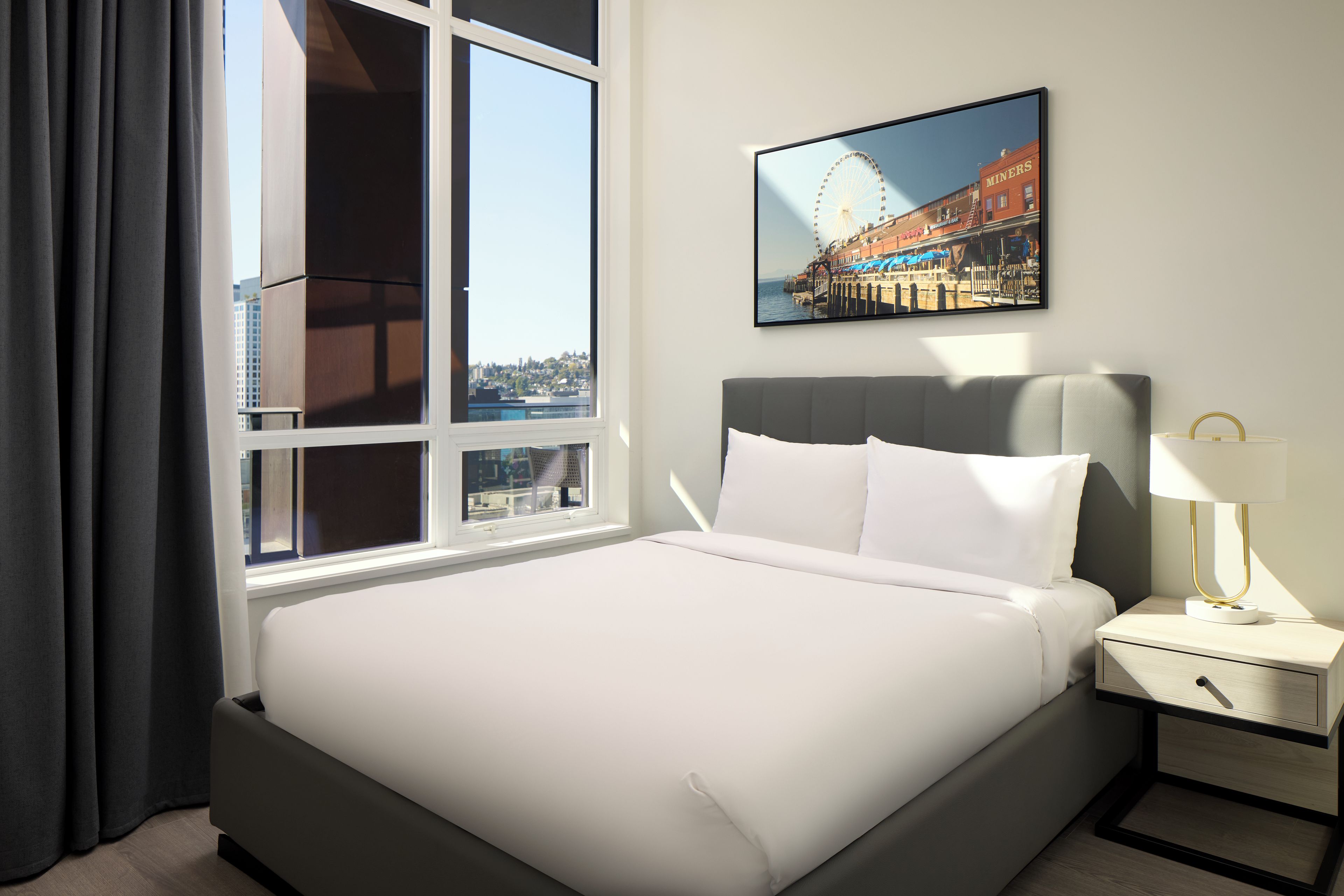 Una habitación de hotel con una cama grande, una mesita de noche con lámpara, una ventana con cortinas y un cuadro en la pared.