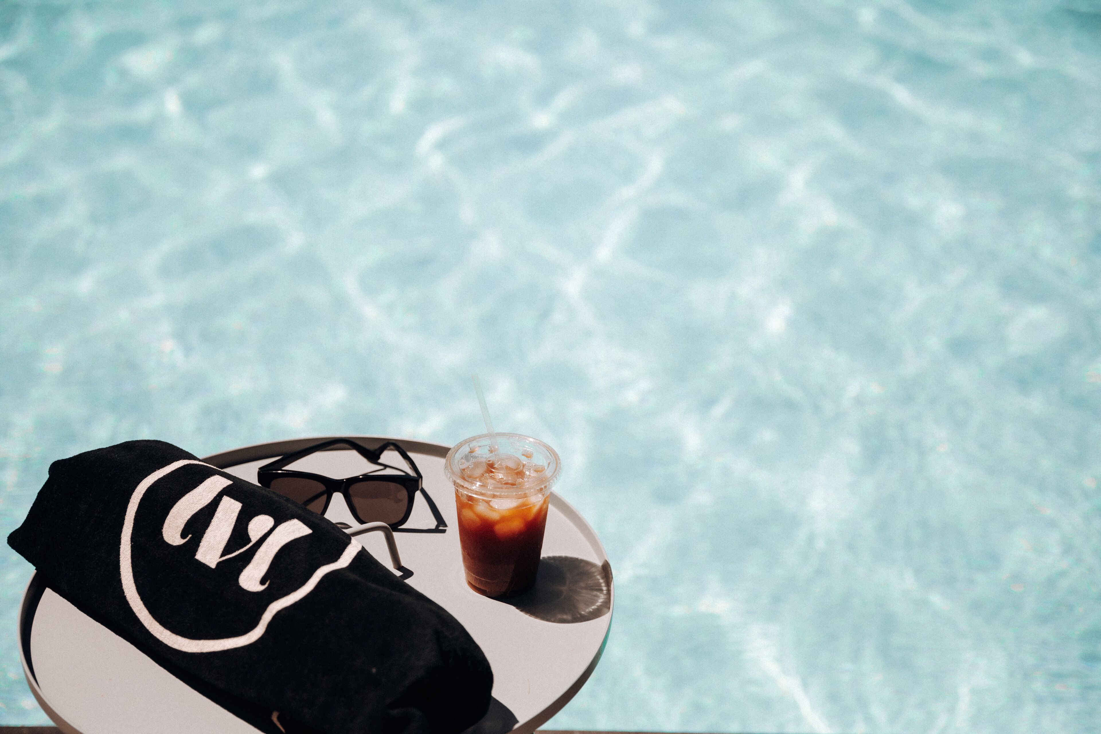 A pair of black and white sneakers, a plastic cup with a straw, and a glass of iced beverage on a round table by a pool.