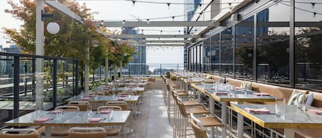 An outdoor dining area with tables and chairs, string lights overhead, and a view of the cityscape.