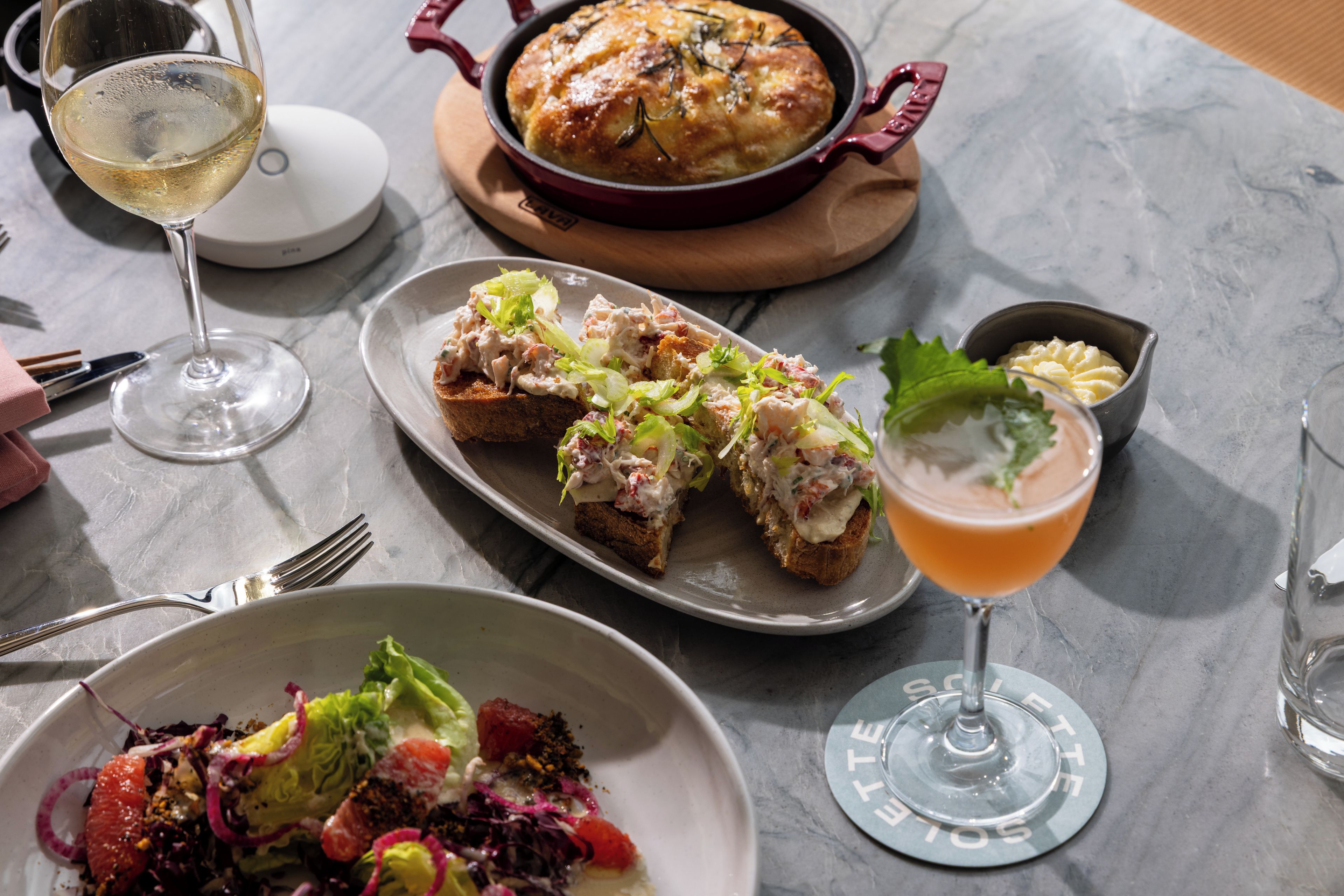 A dining table with a variety of dishes including a salad, a breaded item with a creamy topping, a baked dish in a red pot, and a glass of a pinkish beverage.