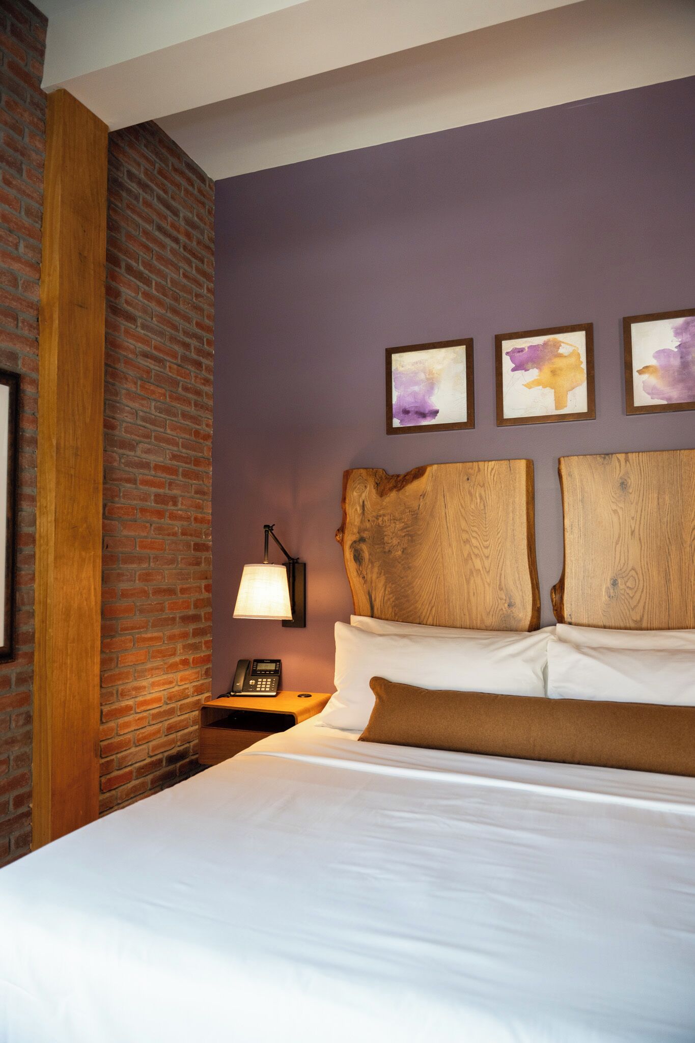 A hotel room with a brick accent wall, a wooden headboard, a white bed, and a nightstand with a lamp.