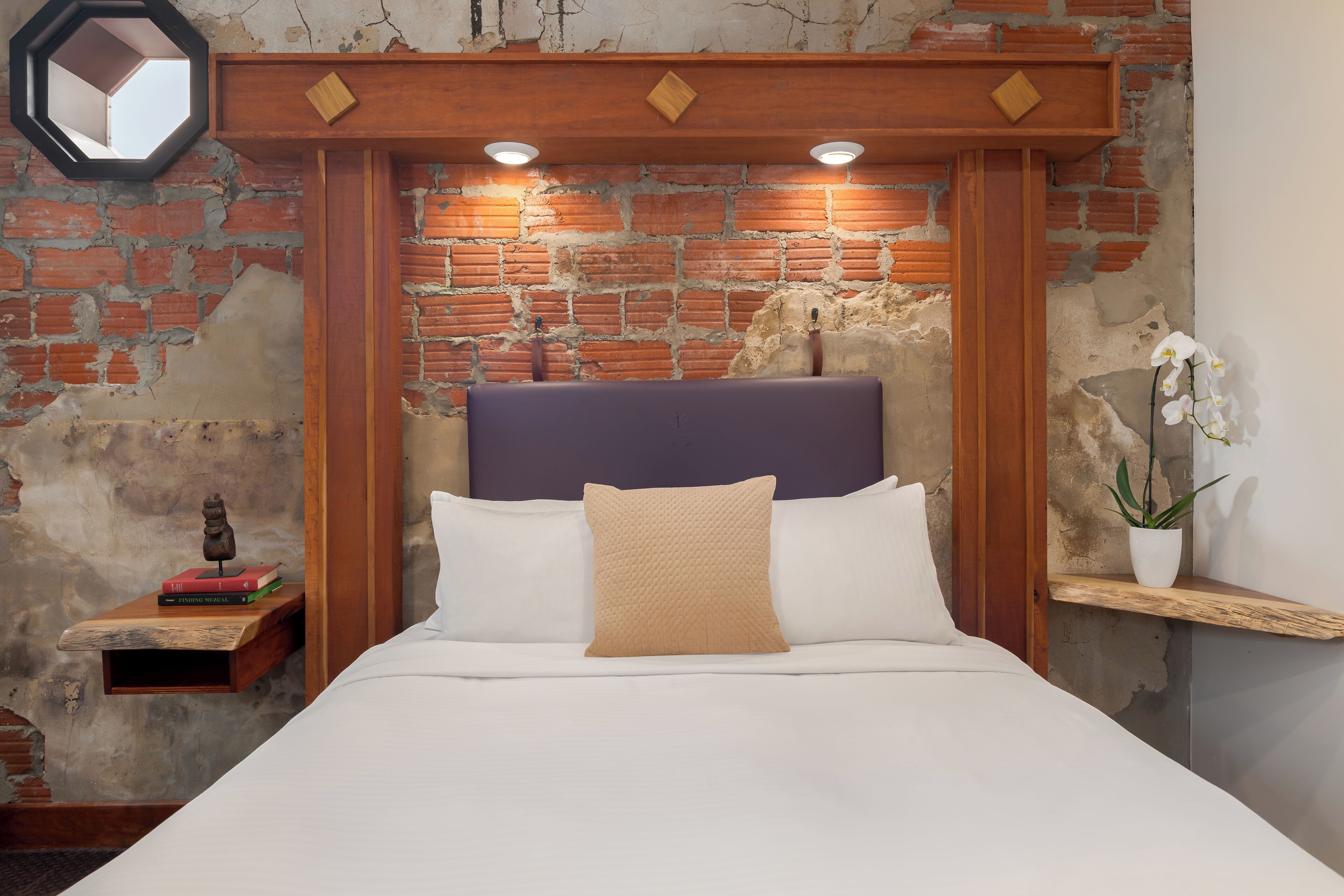 Een bed met een houten hoofdeinde, wit beddengoed en een beige kussen. Er staat een nachtkastje met een kleine plant en een decoratief object.
