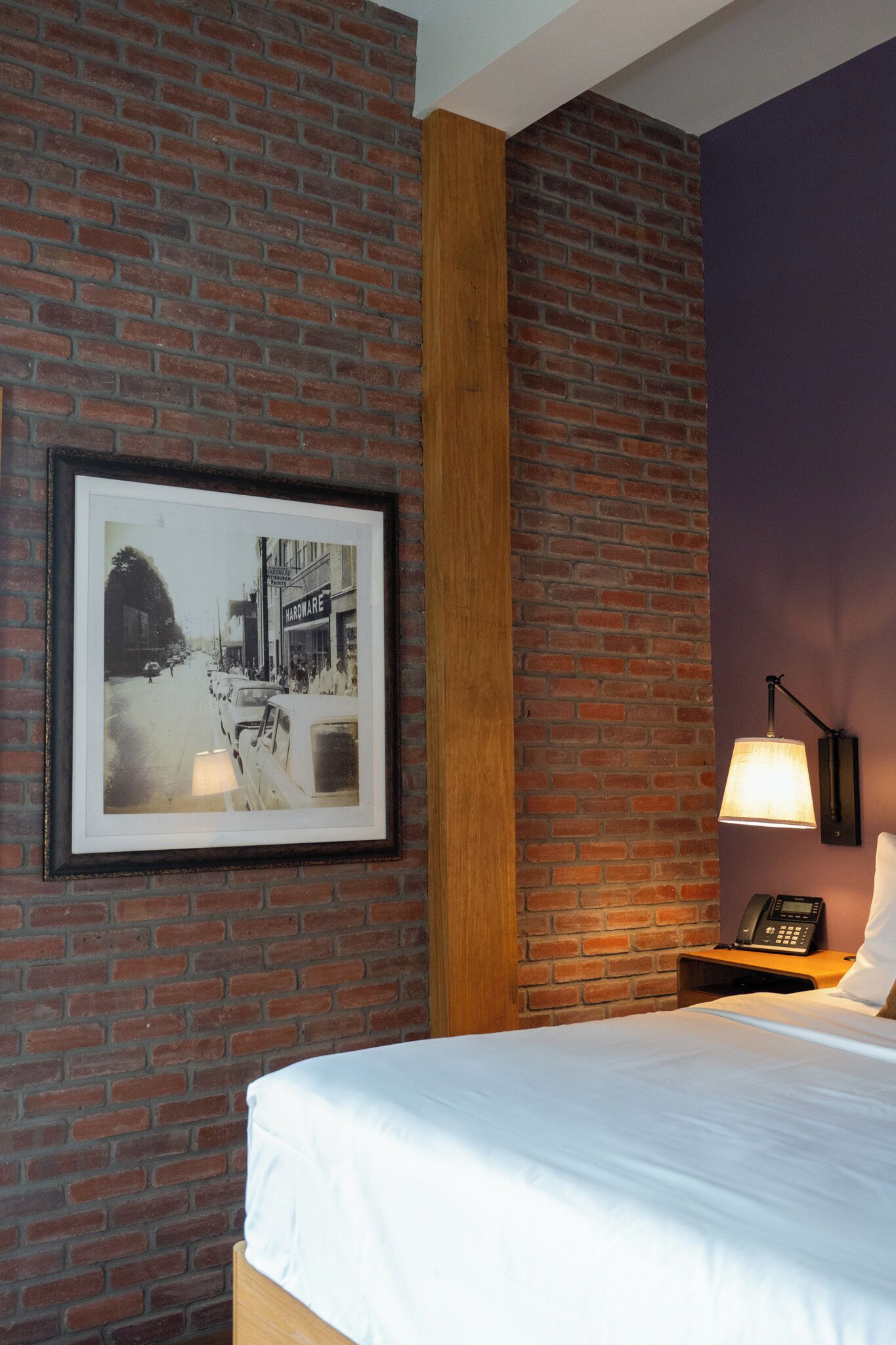 Ein Hotelzimmer mit Backsteinwand, einem gerahmten Bild, einem Bett, einem Nachttisch mit Lampe und einem Telefon.