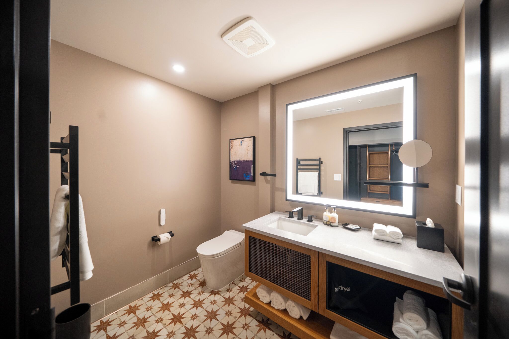 Un baño moderno con un espejo grande, un lavabo blanco y un inodoro. Hay toallas y artículos de aseo en la encimera.