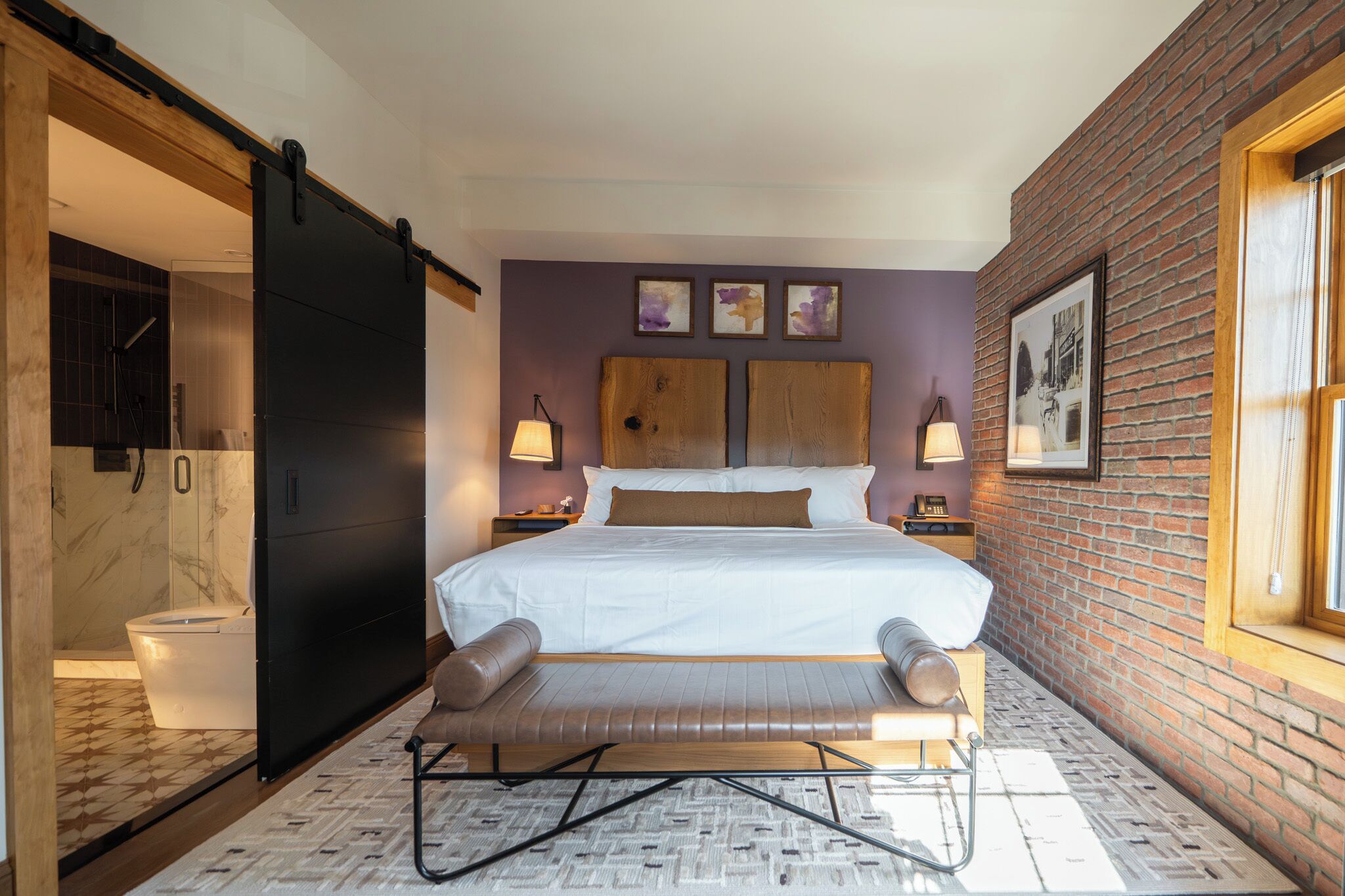A modern bedroom with a large bed, a bench, a sliding door, and a brick wall.