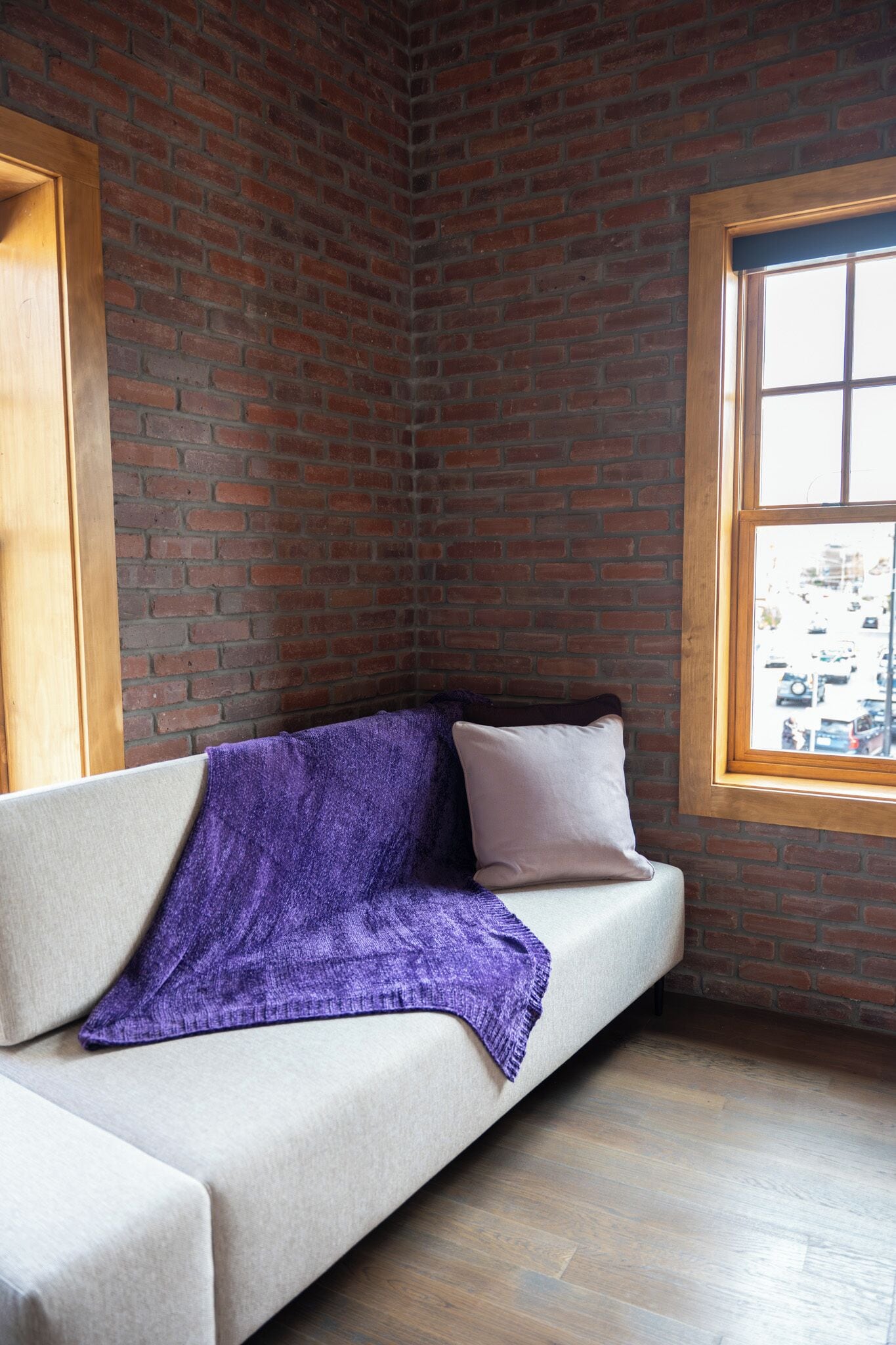 Une pièce d’angle avec un mur en briques, une fenêtre et un canapé recouvert d’un plaid violet.