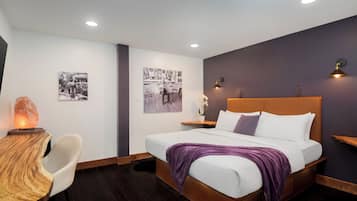 A modern bedroom with a wooden headboard, a white bed, a purple throw blanket, a wooden side table with a salt lamp, and wall art.