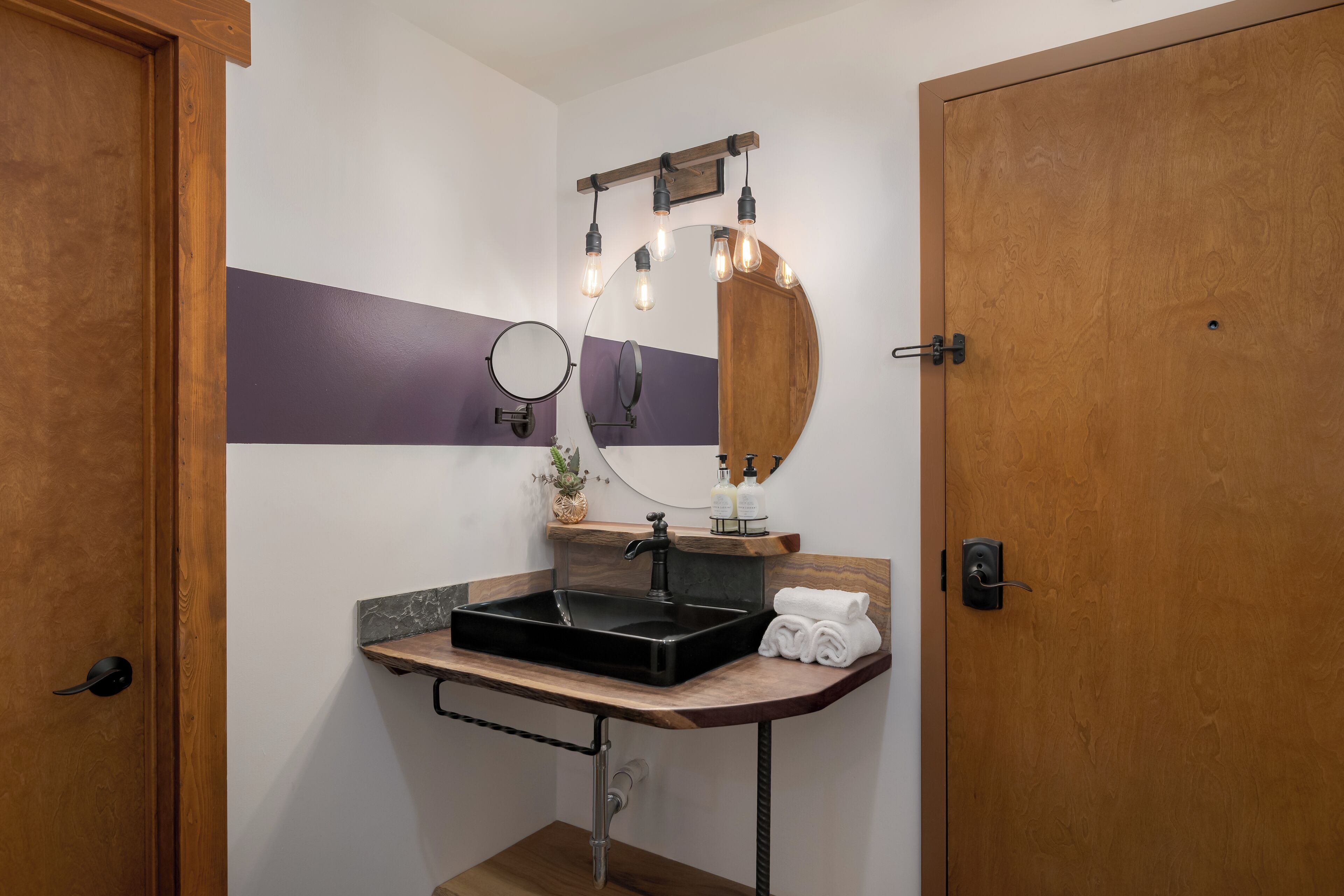 Un baño con tocador de madera, lavamanos negro, espejo con luces y toallero.
