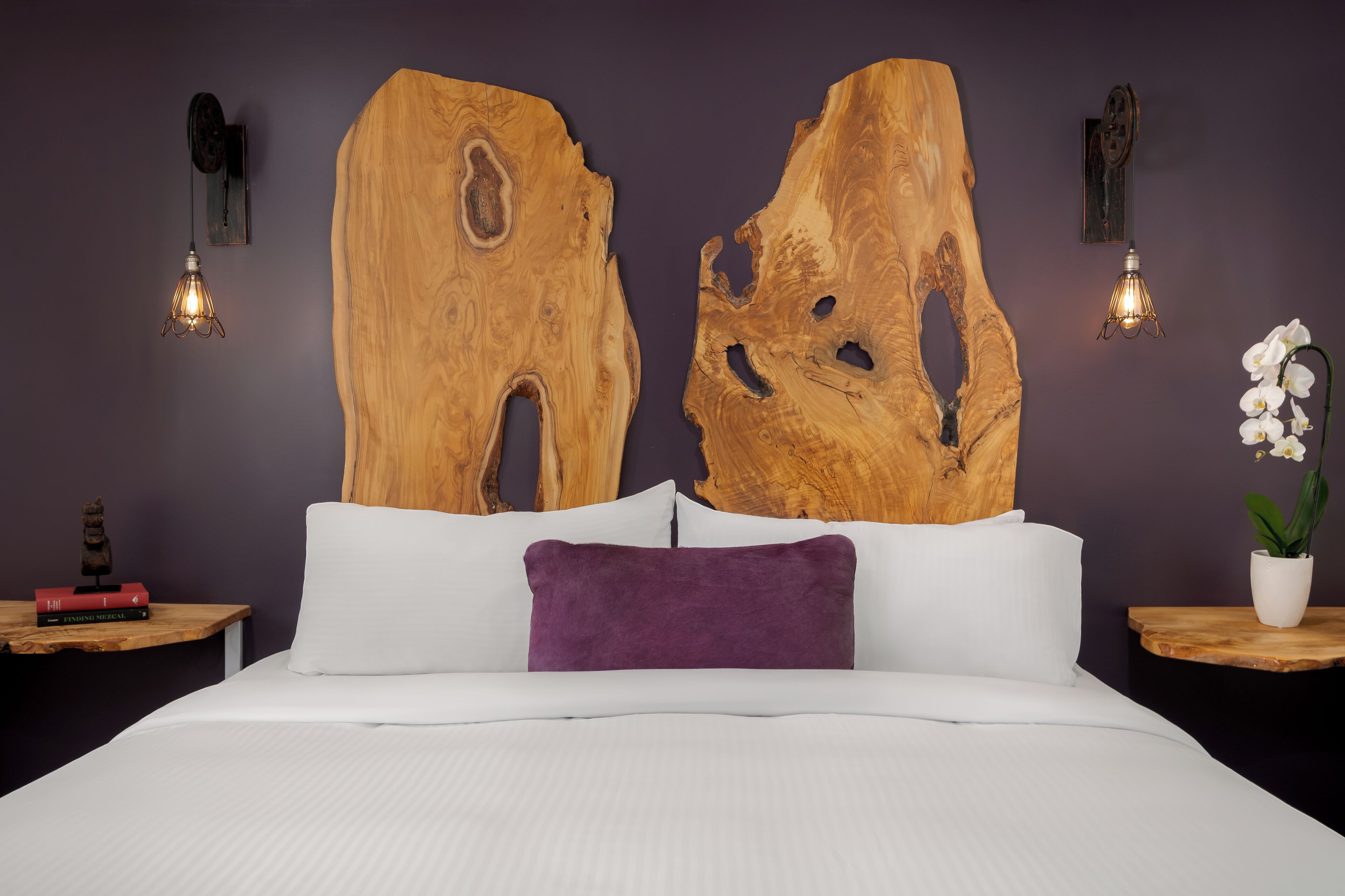 Un dormitorio con una cama grande, dos piezas de arte de madera en la pared, un almohadón blanco, un almohadón púrpura, una pequeña estatua y dos luces fijadas a la pared.