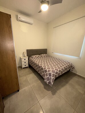 Un dormitorio con una cama, una mesita de noche, una lámpara, un aire acondicionado y una ventana.