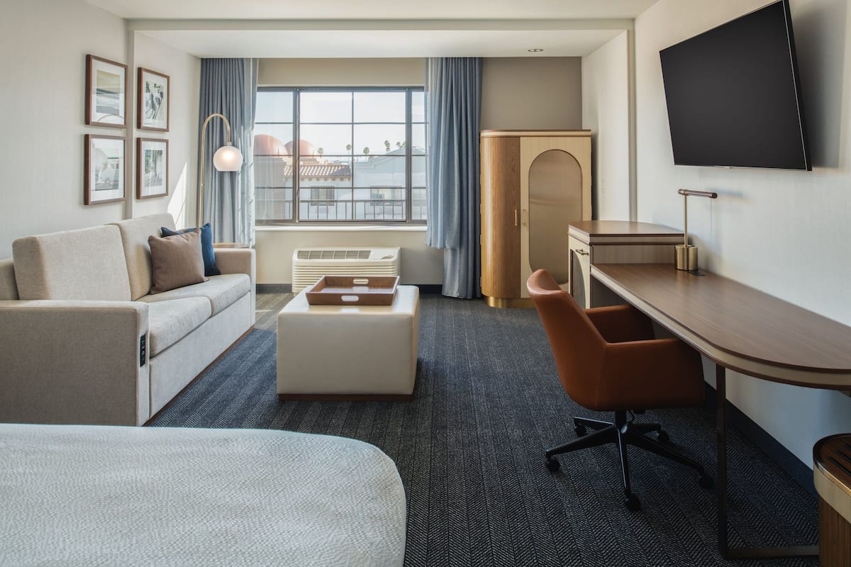 A hotel room with a bed, a sofa, a desk, a chair, a TV, and a window with curtains.