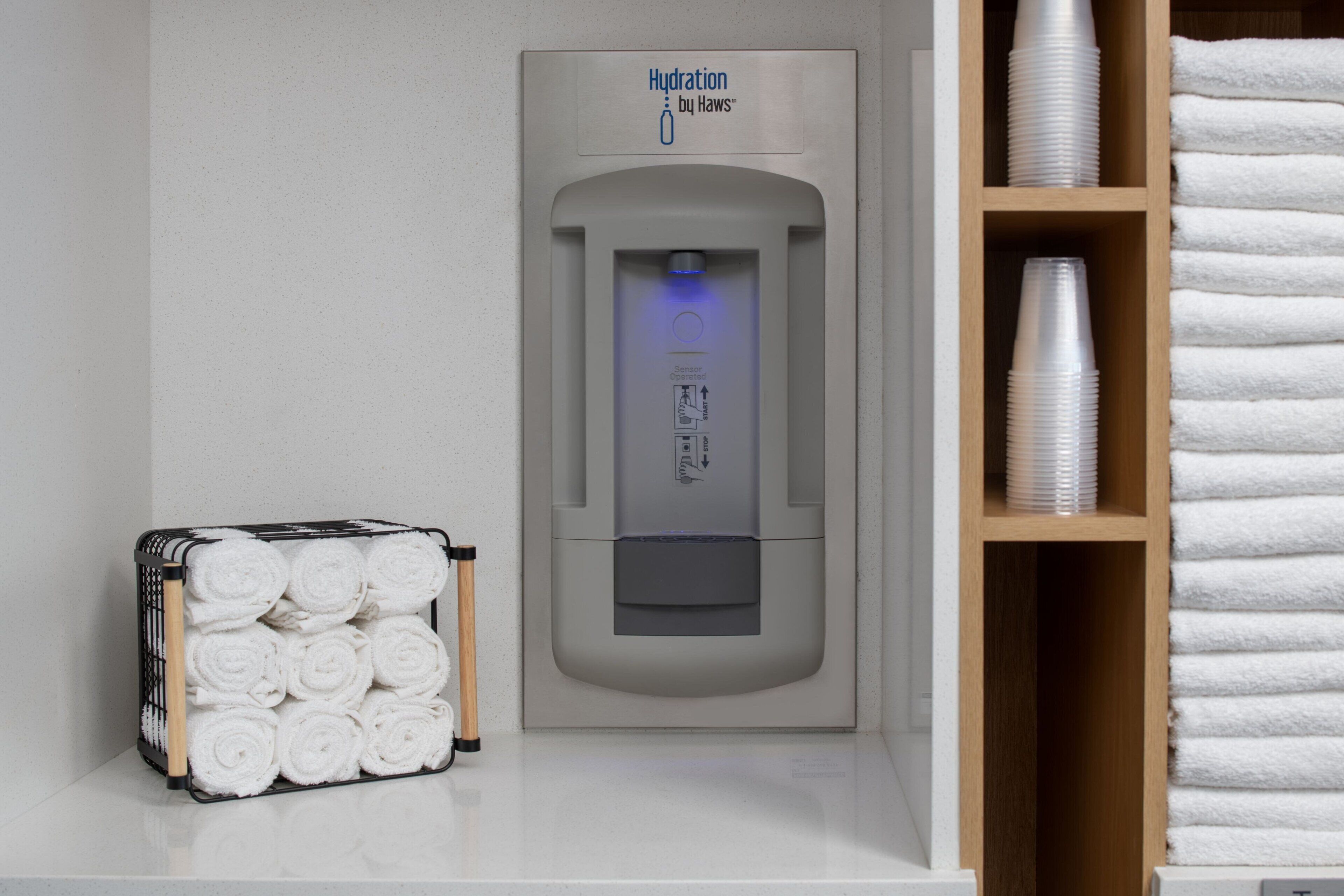 A hydration station with a sensor, a towel rack with neatly rolled towels, and a shelf with stacked paper cups.