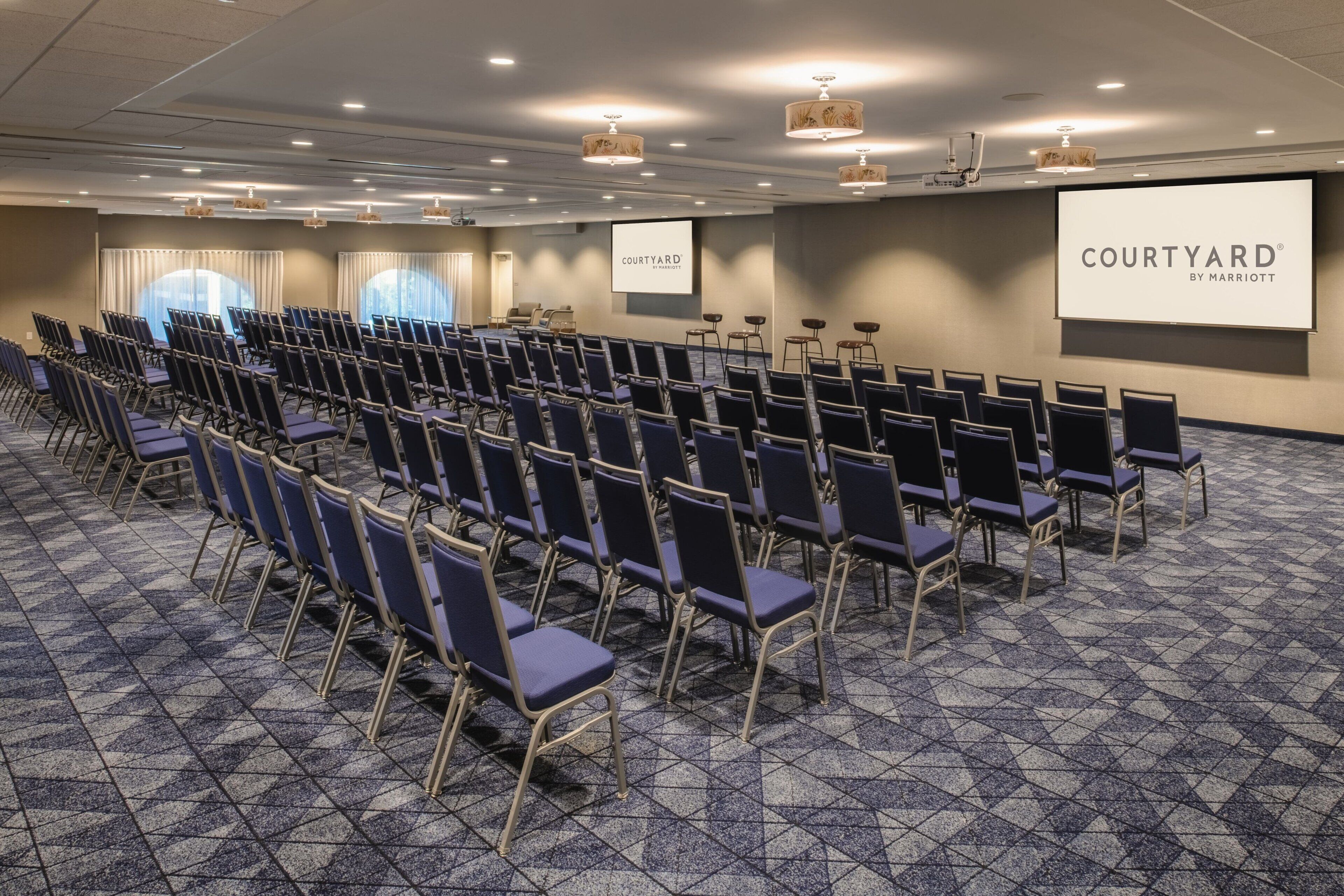 A conference room with rows of chairs, a large screen displaying 'COURTYARD BY MARRIOTT', and a carpeted floor.