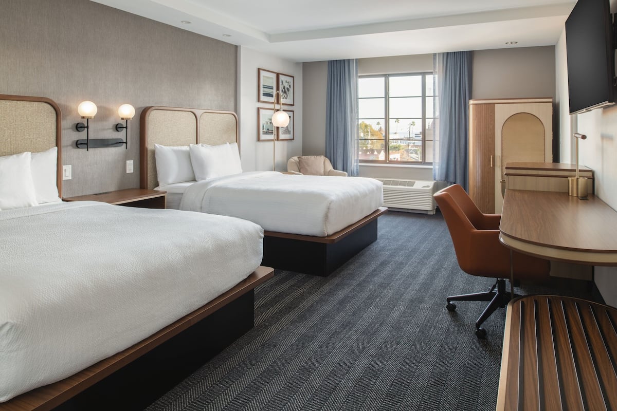 A hotel room with two beds, a desk, a chair, a TV, and a large window with curtains.