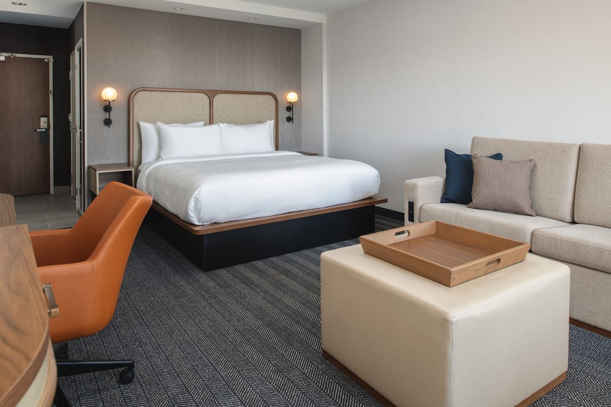 A hotel room with a bed, a sofa, a chair, and a wooden tray on a ottoman.