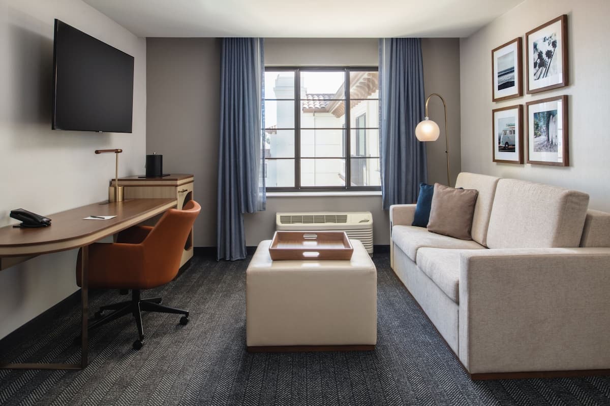 A hotel room with a flat-screen TV, a desk with a chair, a sofa, a coffee table, and a window with curtains.