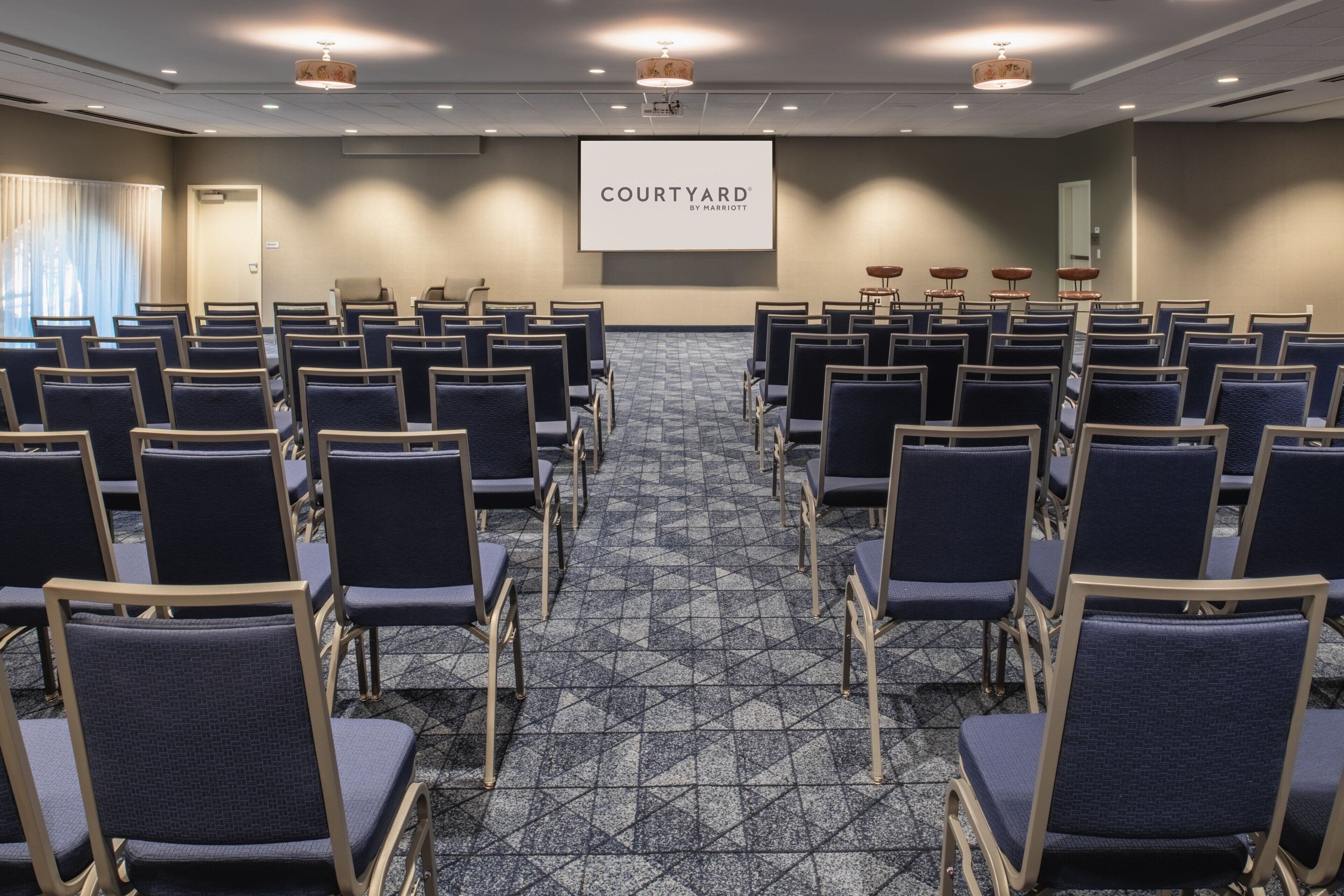 Uma sala de conferências com fileiras de cadeiras, uma tela de projetor exibindo "COURTYARD BY MARRITTON" e piso carpeteado.