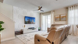 Ein modernes Wohnzimmer mit einem beigefarbenen Ledersofa, einem Flachbildfernseher an einer weißen Wand mit geometrischen Mustern, einem Deckenventilator und einem großen Fenster mit durchsichtigen Vorhängen.