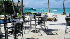 Un espace extérieur aménagé avec des tables et des chaises, offrant une vue sur une plage où l’on peut apercevoir des bateaux et des palmiers.